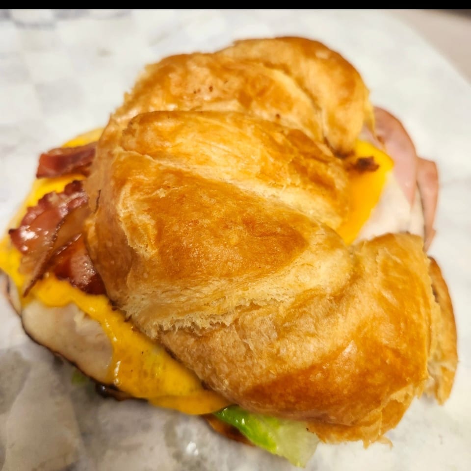 A croissant sandwich with bacon, cheese, ham, and lettuce on white paper.